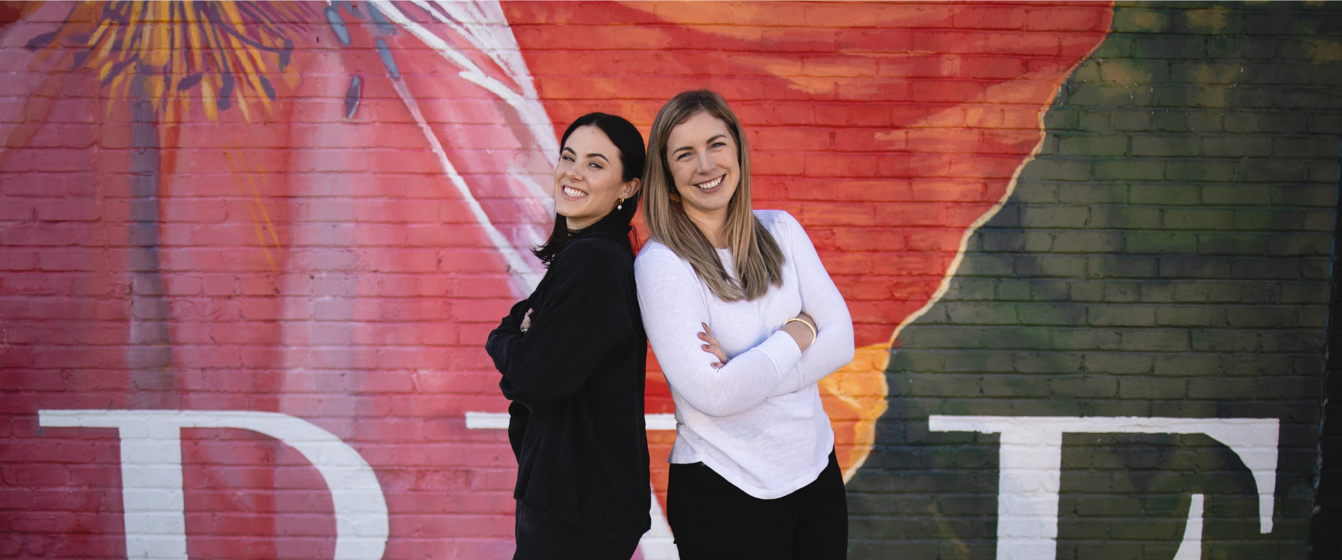 Two women standing back to back smiling with arms cross in front of a mural.